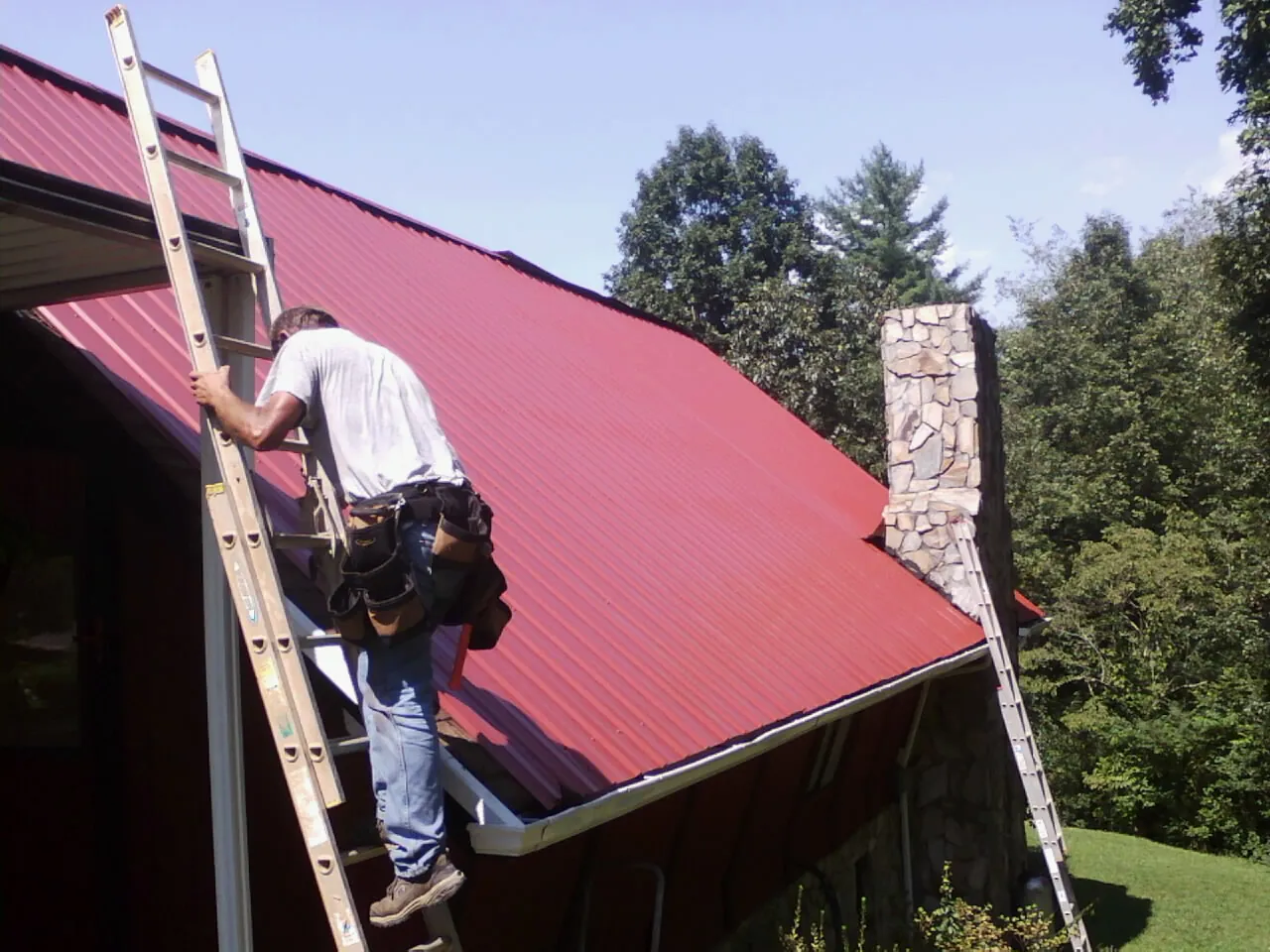 Roofer on a ladder beside a red metal roof and stone chimney — Soffit & Fascia Repair by Roofing Brothers in Thrall